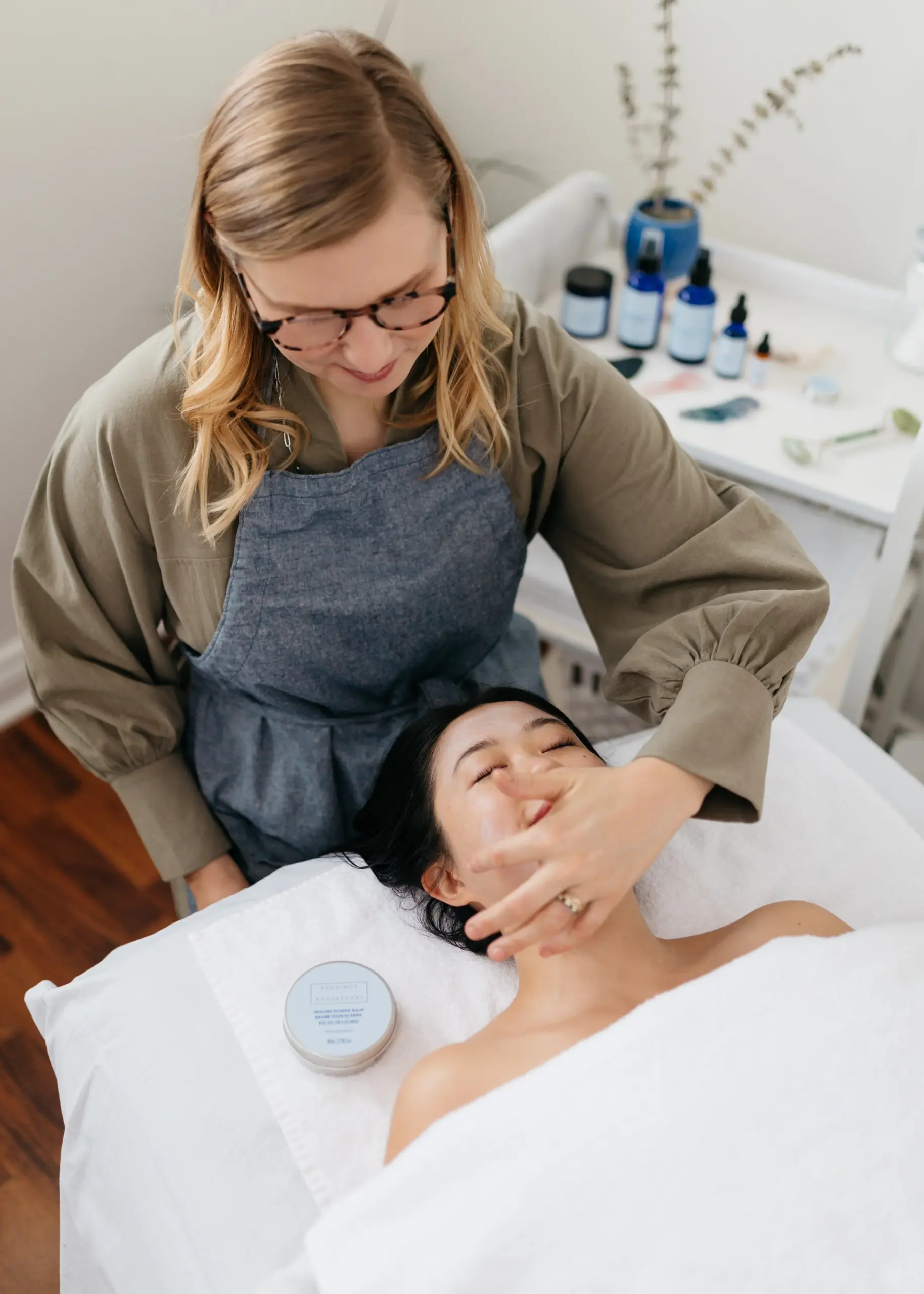 Photo of an in-progress facial session, Julie is applying product to a woman lying on a table in the studio.