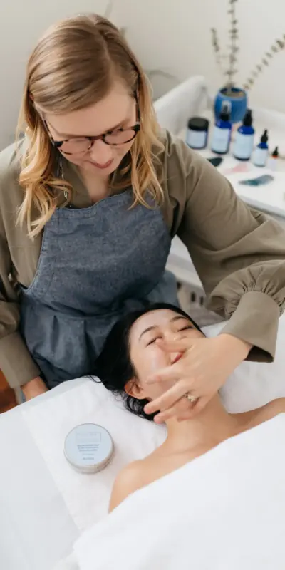 Photo of an in-progress facial session, Julie is applying product to a woman lying on a table in the studio.